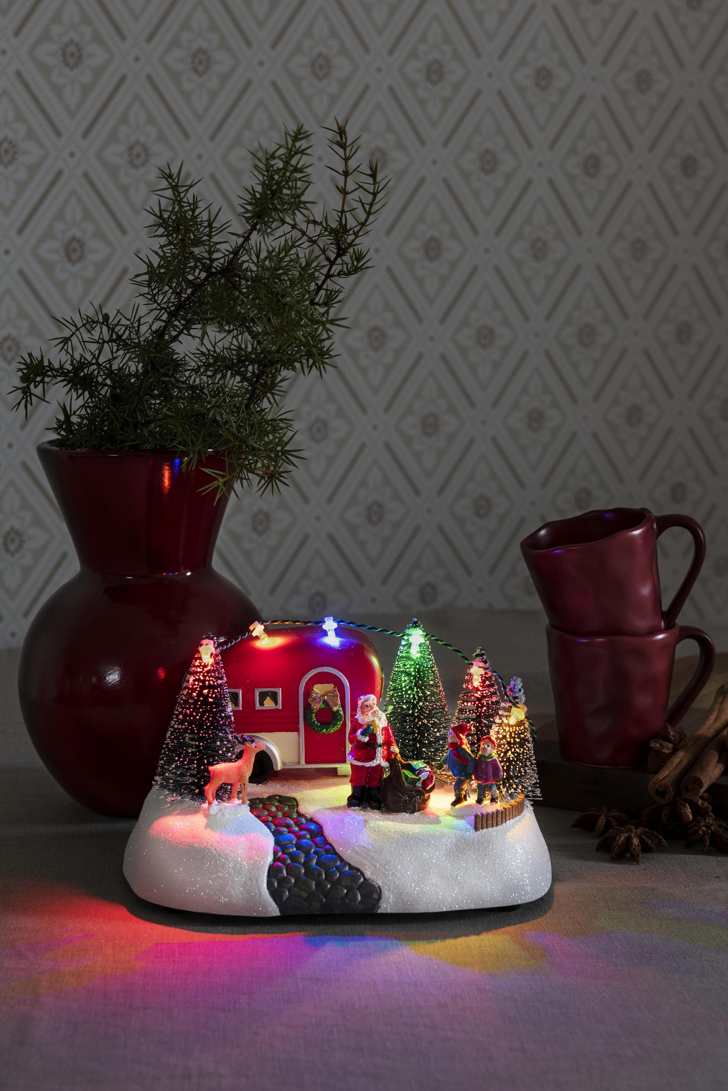 Weihnachtsszene: beleuchteter Wohnwagen, Weihnachtsmann, Bäume, Schnee, Rentiere, rote Vase, Tassen, Zimt.
