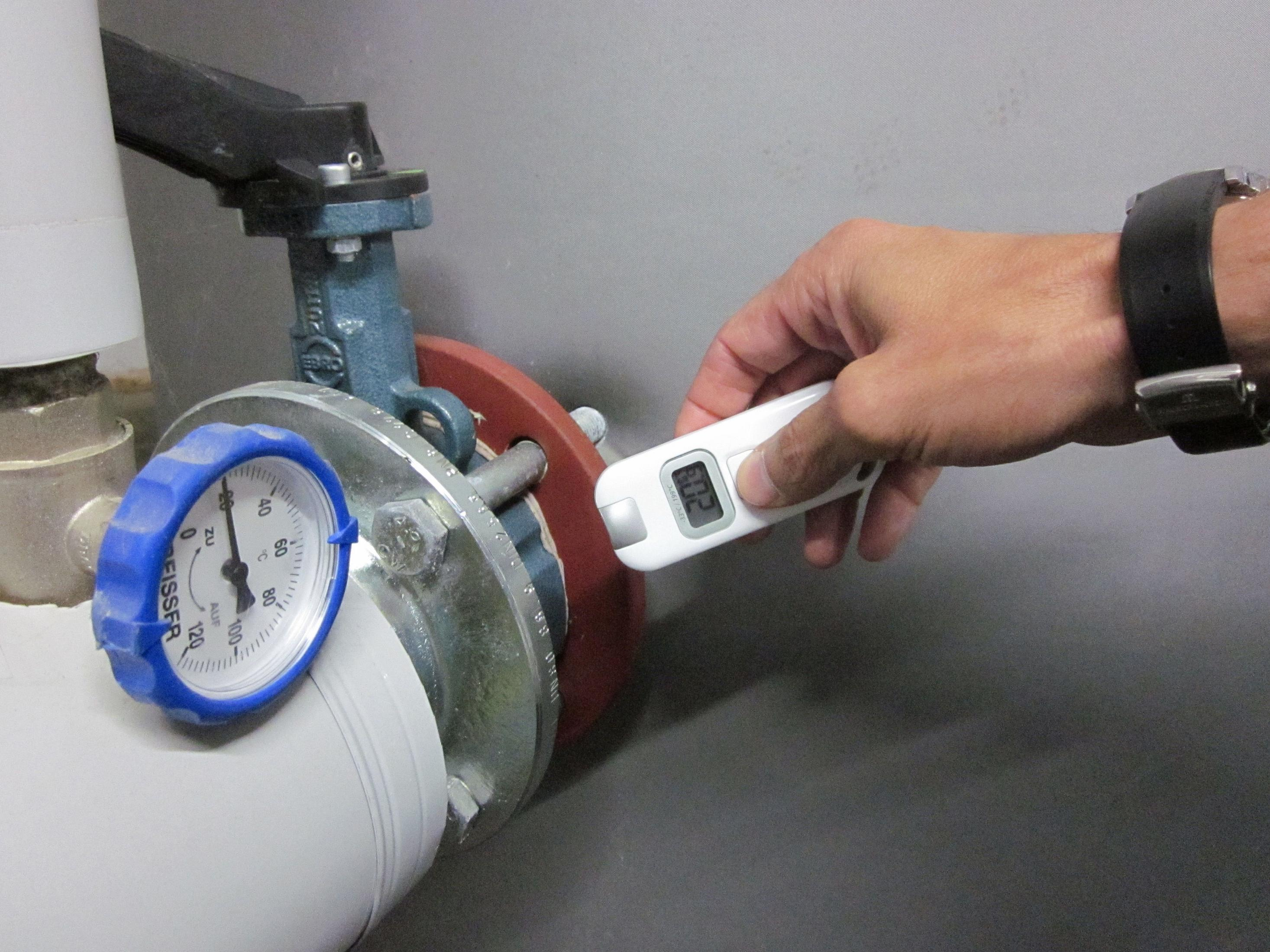 Eine Person hält ein digitales Thermometer in der Nähe eines Rohrs mit Messgeräten und Armaturen in einer industriellen Umgebung.