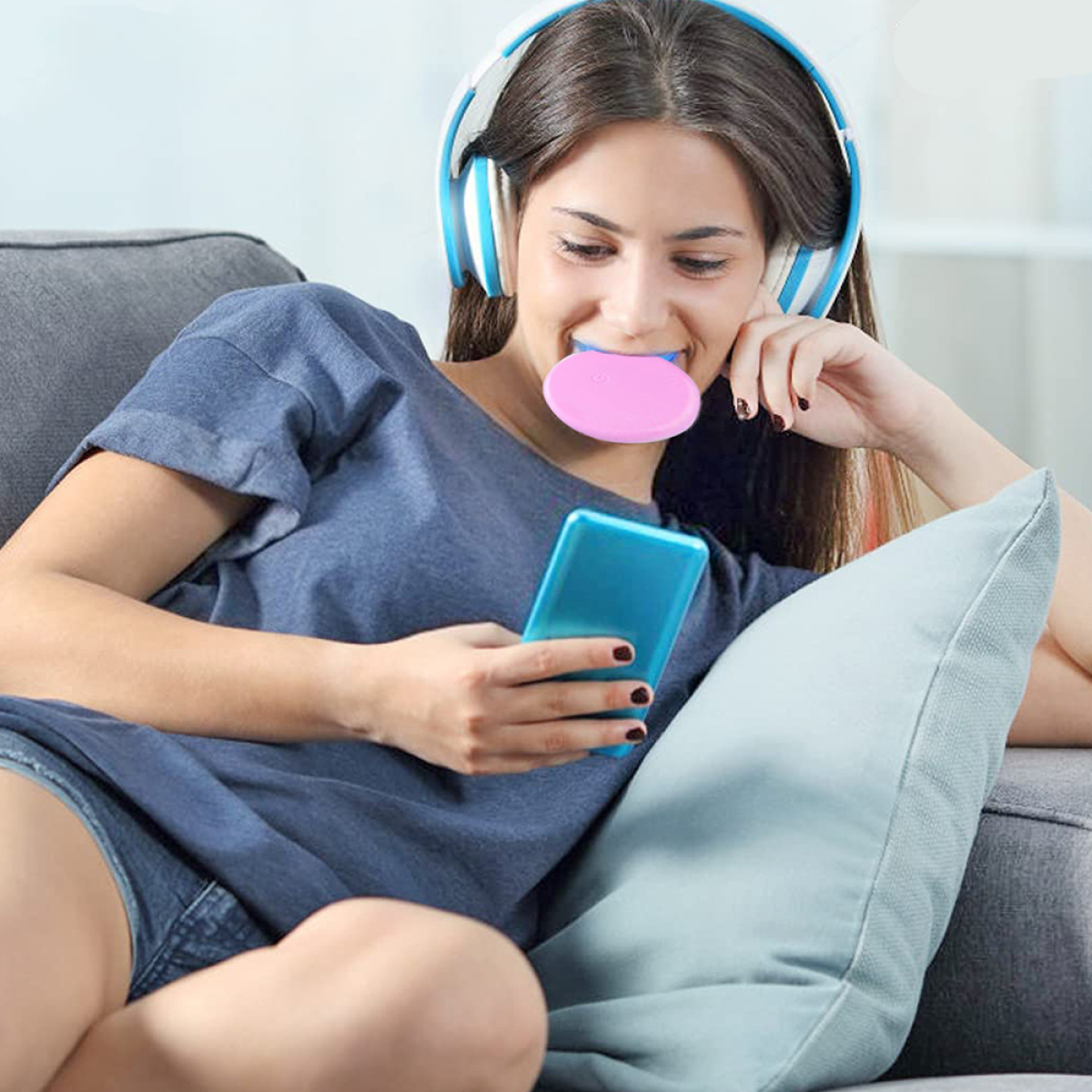 Frau auf einem Sofa, die Kopfhörer trägt, ein Telefon hält und ein Zahnaufhellungsgerät im Mund benutzt.