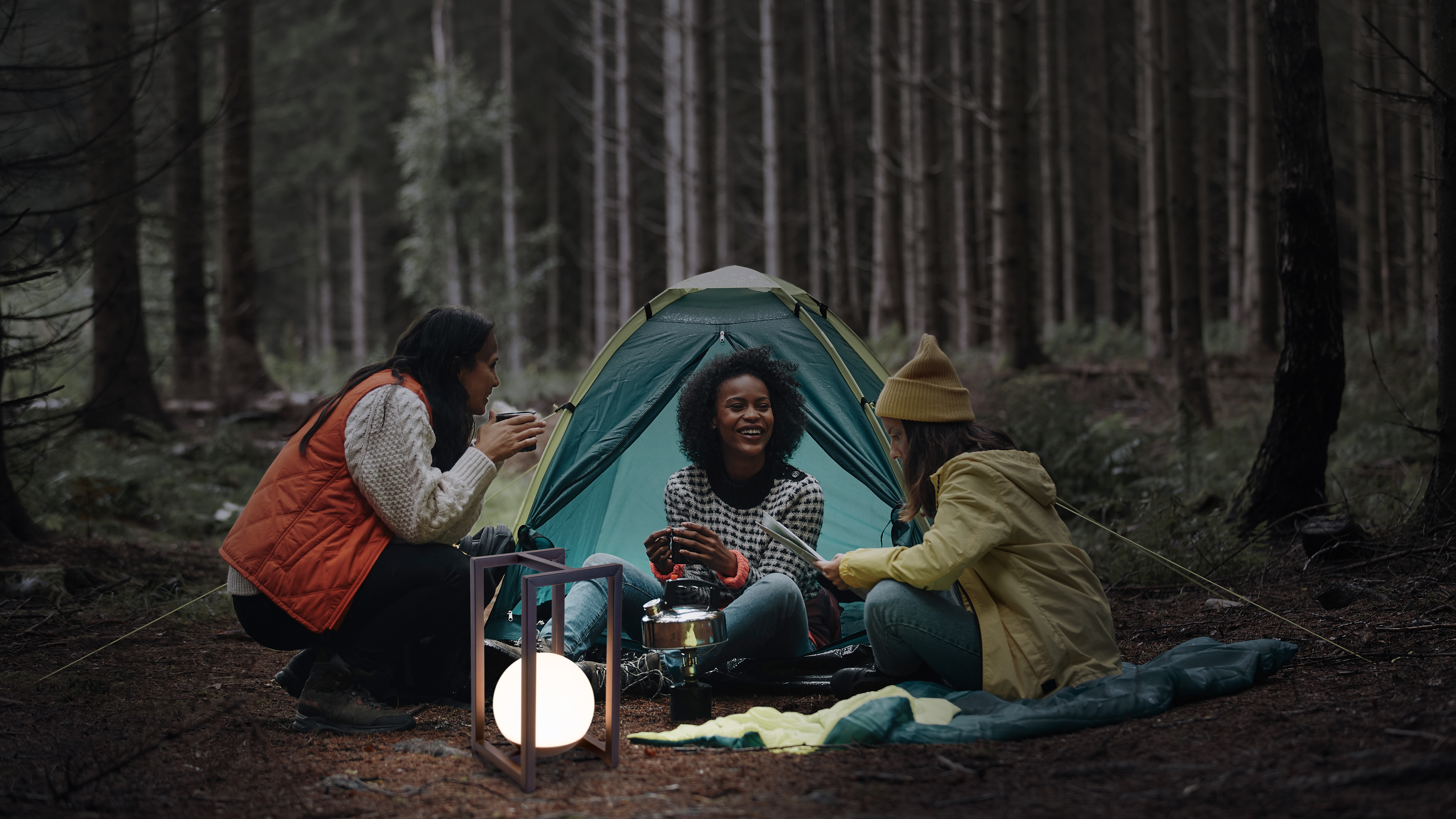Drei Frauen um ein Zelt im Wald, eine hält einen Becher.