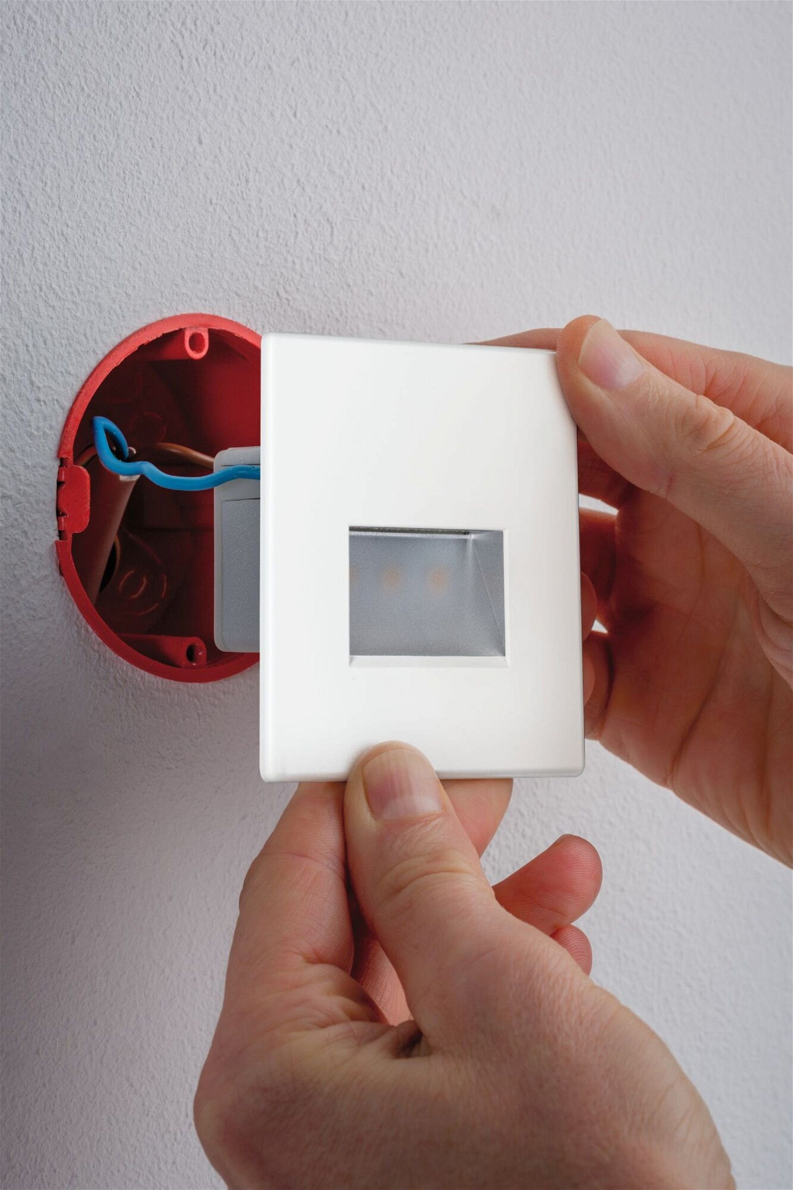 Ręce trzymają białą oprawę oświetleniową, w tle czerwoną skrzynkę elektryczną.