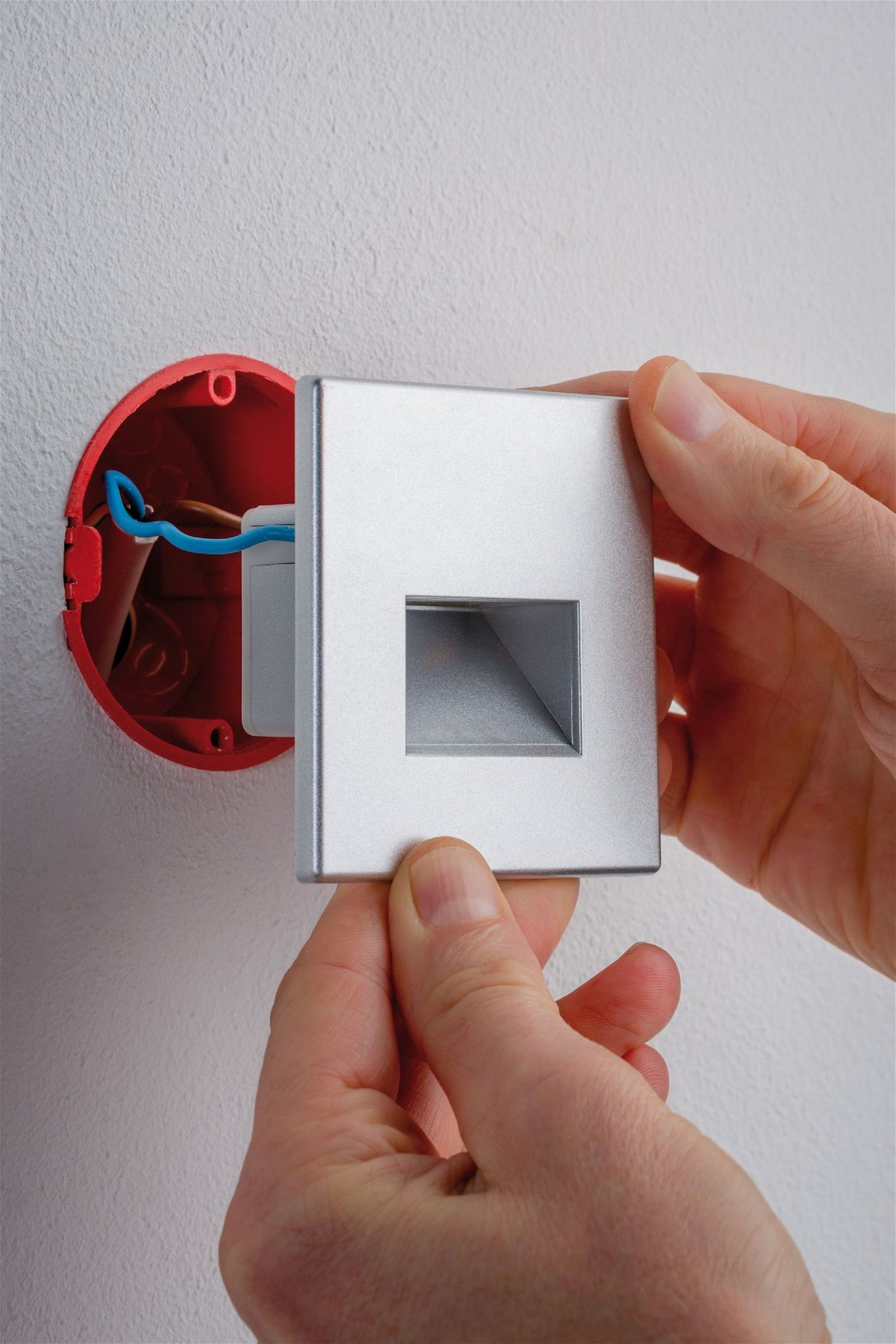 Dłonie instalujące srebrną płytkę ścienną w czerwonej puszce elektrycznej.