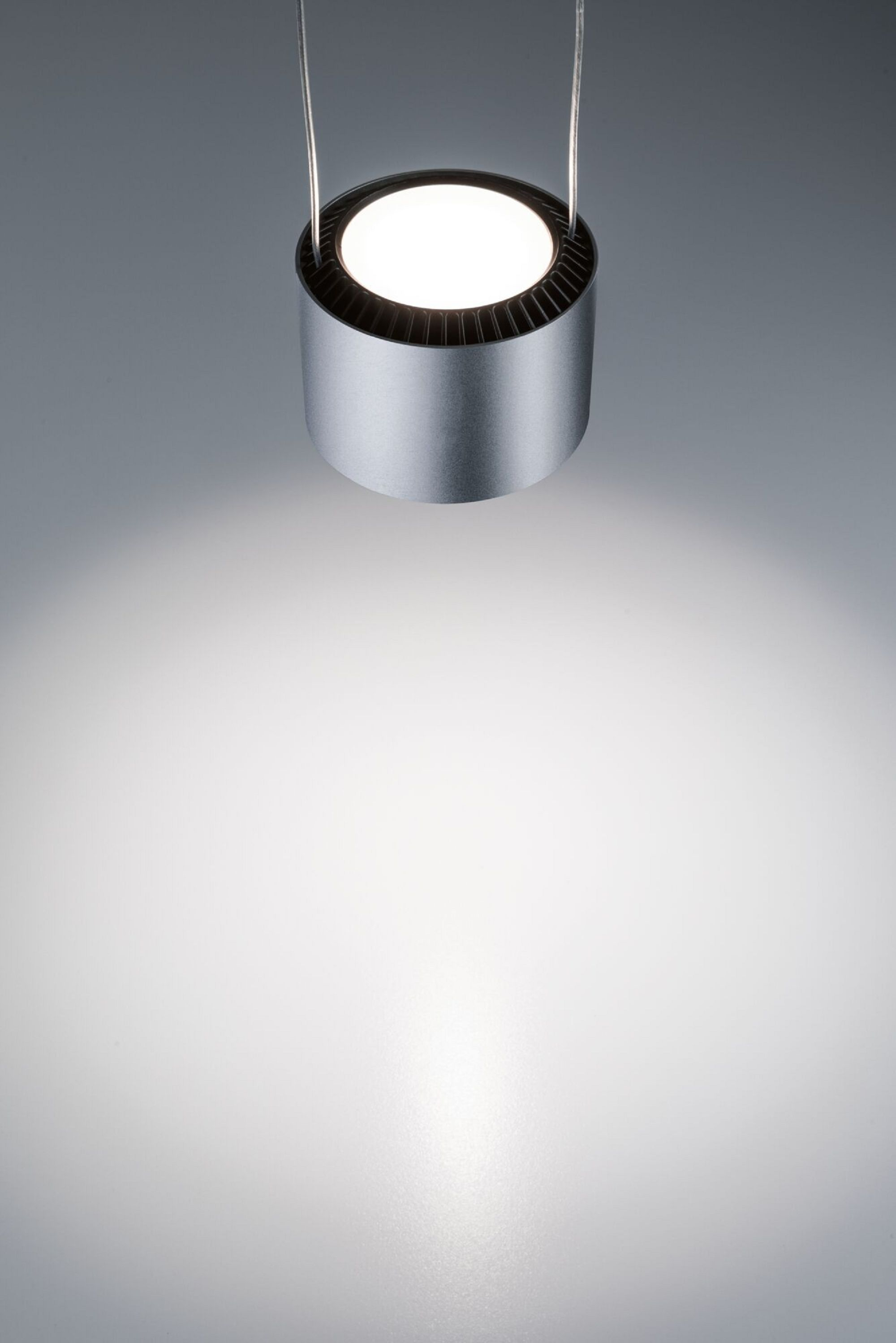 Eine silberne zylindrische Leuchte, die an zwei Drähten hängt. Das Licht ist an und scheint nach unten.
