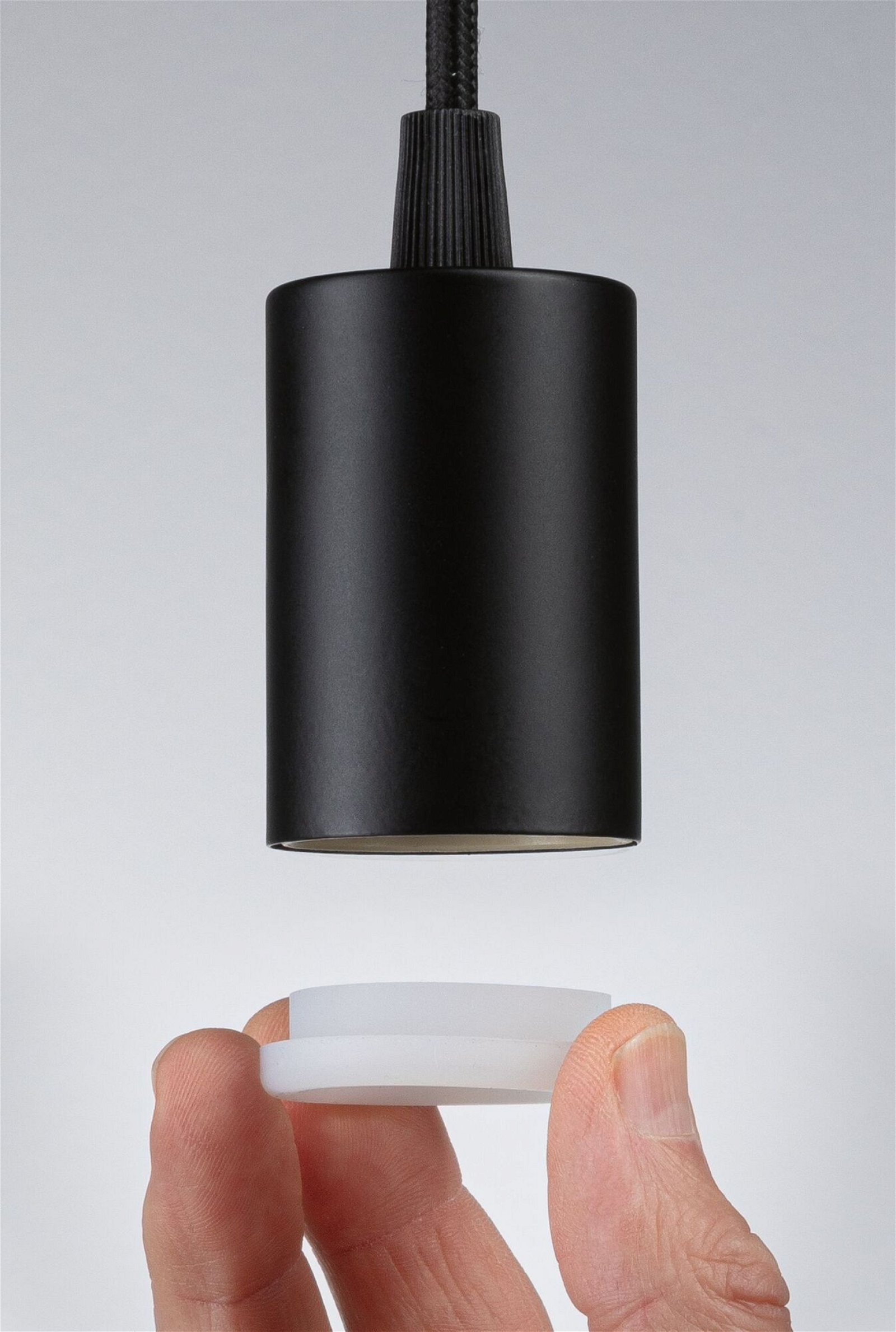 Czarna lampa wisząca z ręką trzymającą biały element, na jasnym tle.