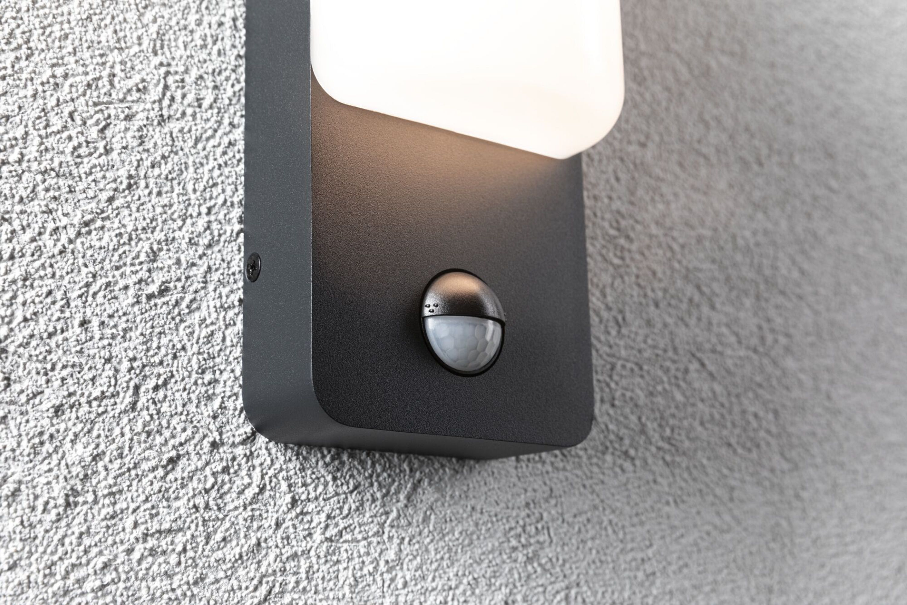 Zewnętrzna lampa ścienna z czujnikiem ruchu na teksturowanej szarej ścianie.