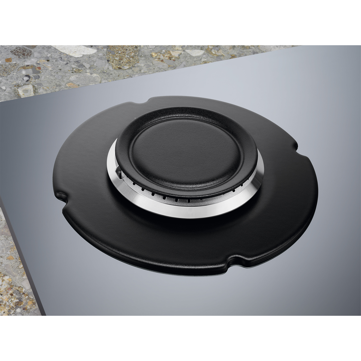 Zbliżenie palnika kuchenki gazowej. Jest czarny ze srebrnym pierścieniem i znajduje się na szarej, wzorzystej powierzchni.