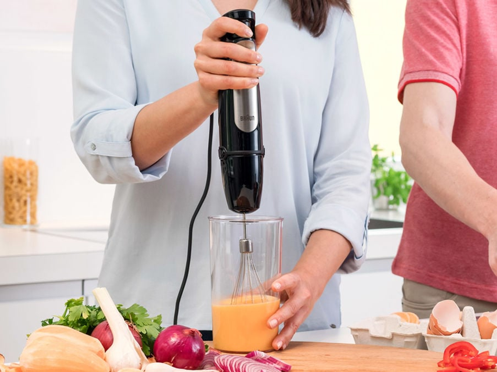 Bir kadın, cam bir kapta yumurtaları karıştırmak için bir Braun el blenderi kullanıyor. Başka bir kişi de var.