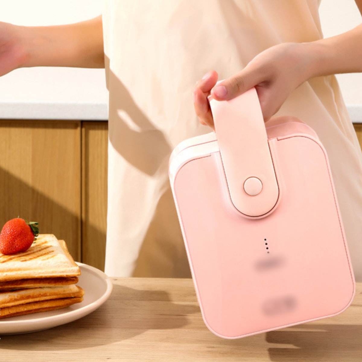 Eine Person hält einen rosa Sandwichmaker neben einem Teller mit Sandwiches und einer Erdbeere.