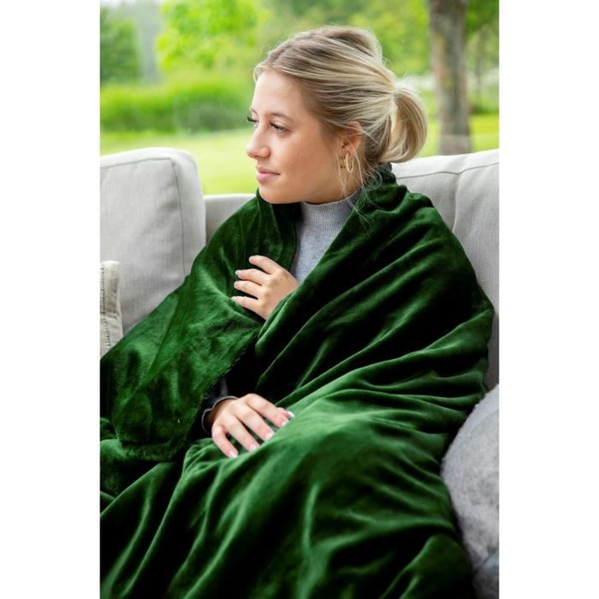 Frau in grüne Decke gehüllt, sitzt auf grauem Sofa, Hintergrund im Freien.