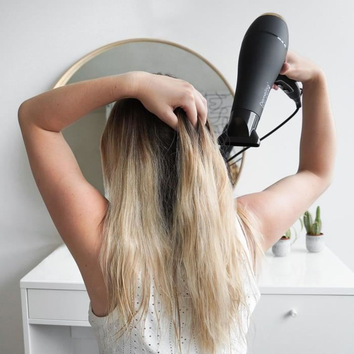 Donna che si asciuga i capelli biondi. Specchio e cassettiera sullo sfondo. Vista posteriore.