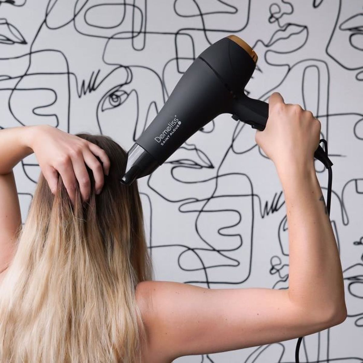 Donna che usa un asciugacapelli nero, asciugando i capelli. Muro bianco a fantasia sullo sfondo.
