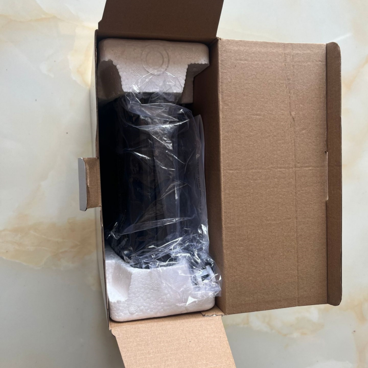 Eine geöffnete Pappschachtel, die ein schwarzes Objekt zeigt, das in Kunststoff und Styropor verpackt ist.