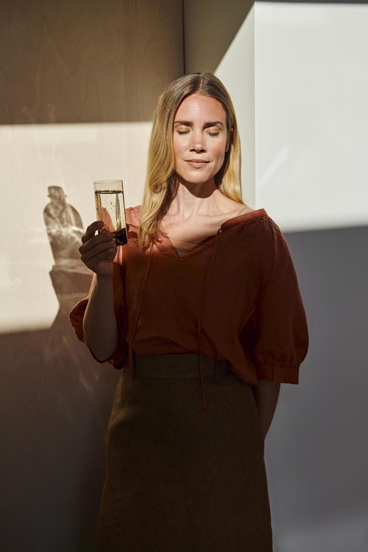 Donna con i capelli biondi tiene un bicchiere d'acqua. Luce solare e ombre sul muro.