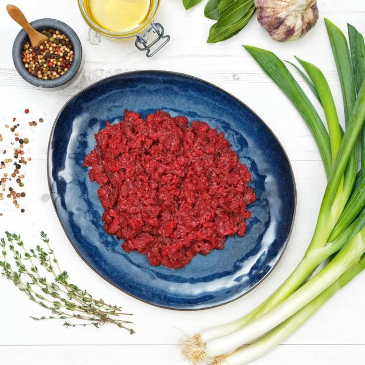 Surowe czerwone mięso na niebieskim talerzu, otoczone ziołami, czosnkiem i szczypiorkiem.