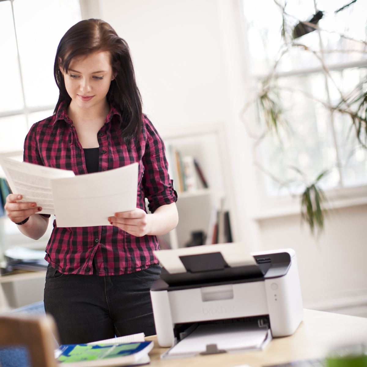 Kobieta stoi przy biurku, czytając papiery, z drukarką na biurku. Tło to pokój z oknem.