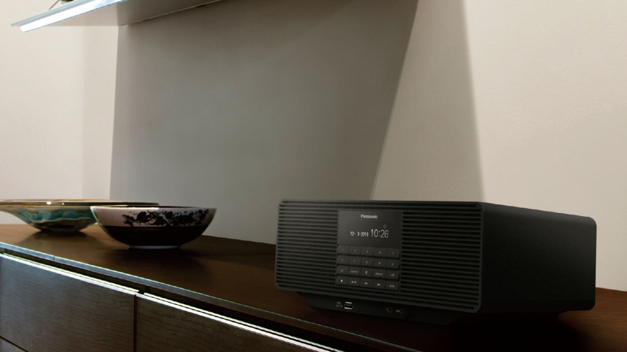 Czarny radioodtwarzacz Panasonic na drewnianej półce z dwiema miskami i białą ścianą w tle.