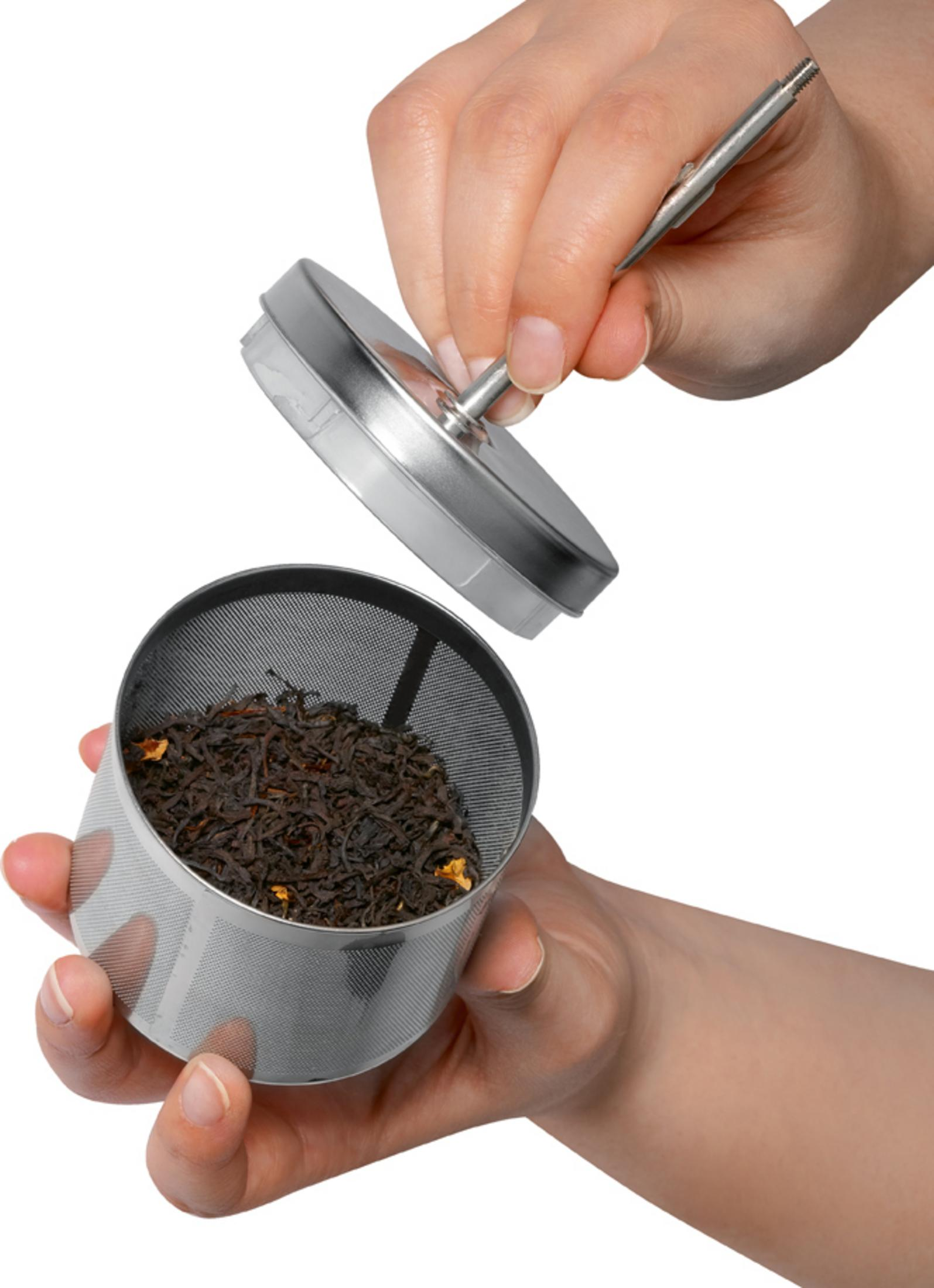 Ręka trzyma zaparzacz do herbaty z liśćmi herbaty. Druga ręka podnosi pokrywkę.