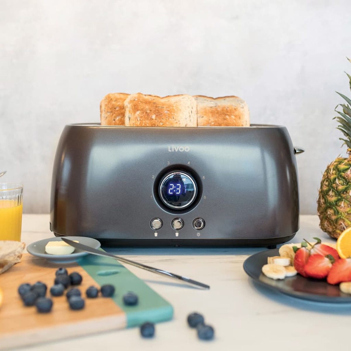 Ein grauer Toaster mit Toast und digitaler Uhranzeige, sowie Obst und Butter.