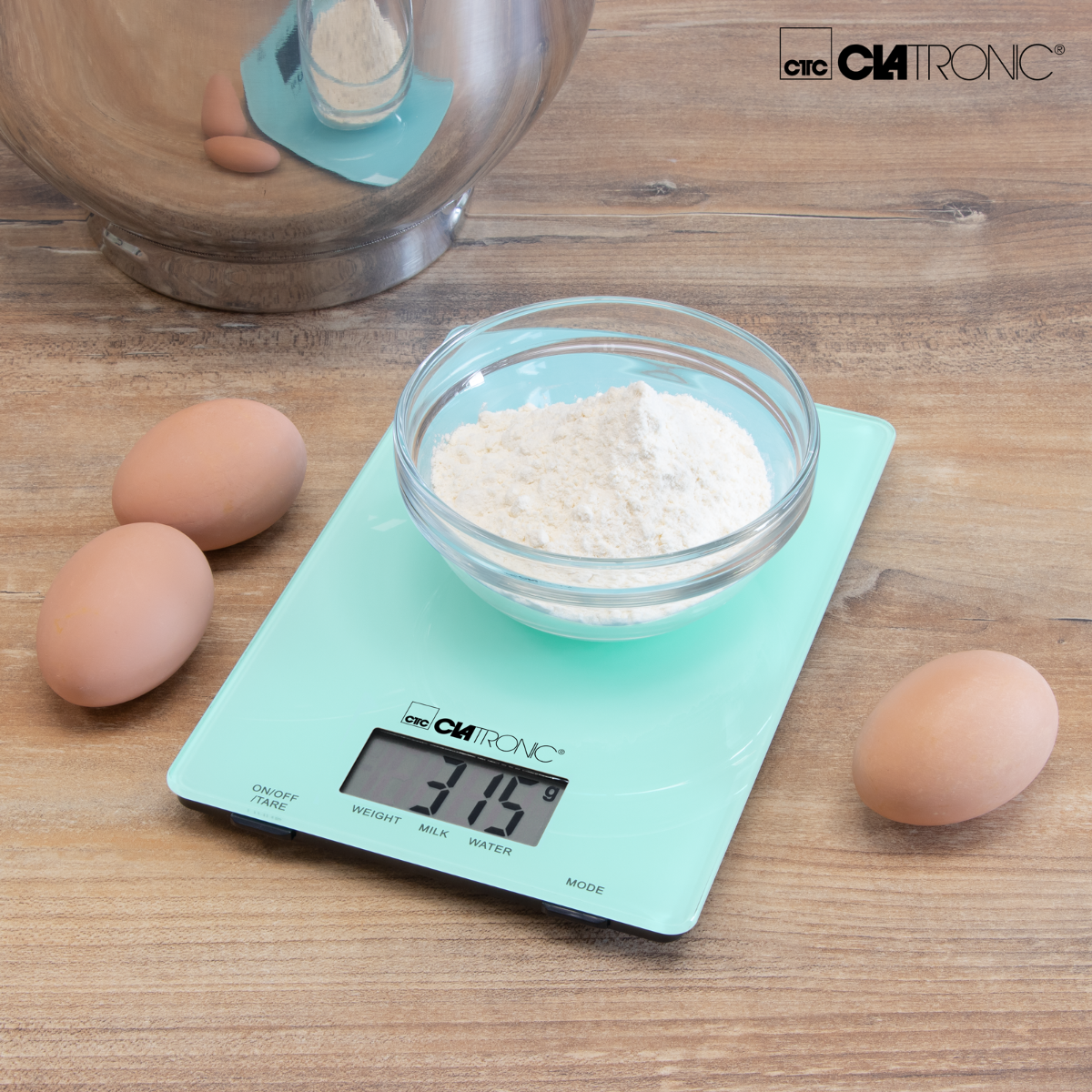 Una bilancia da cucina digitale con farina in una ciotola di vetro, uova e una ciotola per mescolare.
