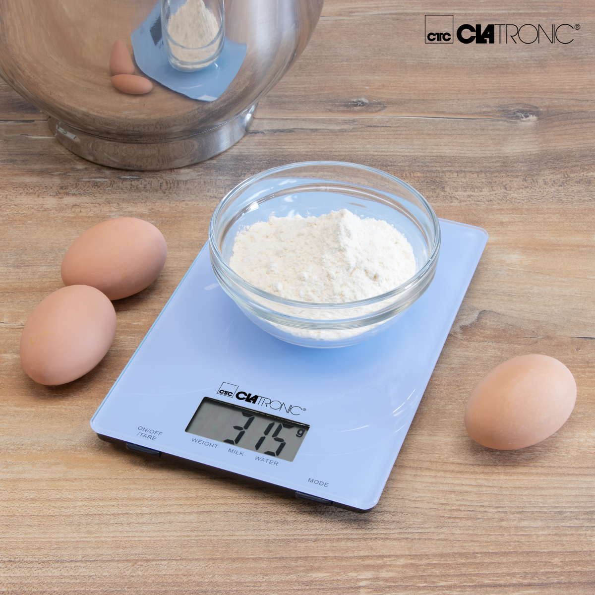 Niebieska cyfrowa waga kuchenna z jajkami i miską mąki. Waga pokazuje 315g.