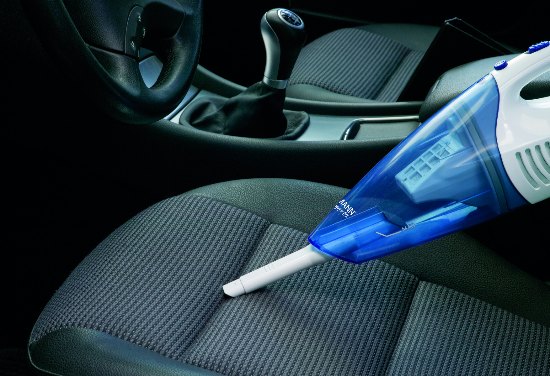 Osoba używająca odkurzacza ręcznego na siedzeniu samochodowym. Odkurzacz jest niebiesko-biały.