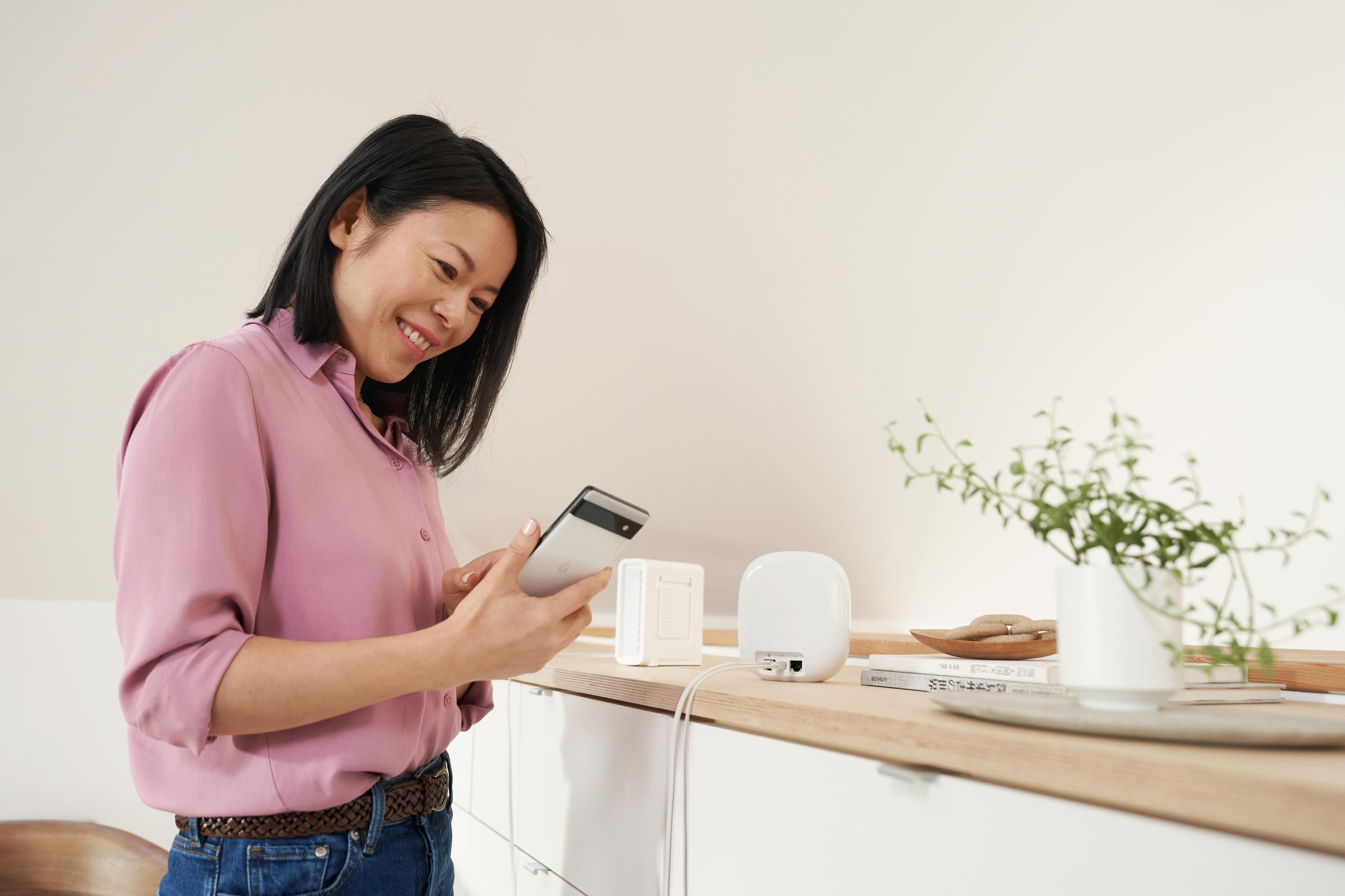 Frau hält Telefon und lächelt. Auf dem Regal befinden sich ein weißes Gerät und ein Router. Pflanzen in einer Vase.