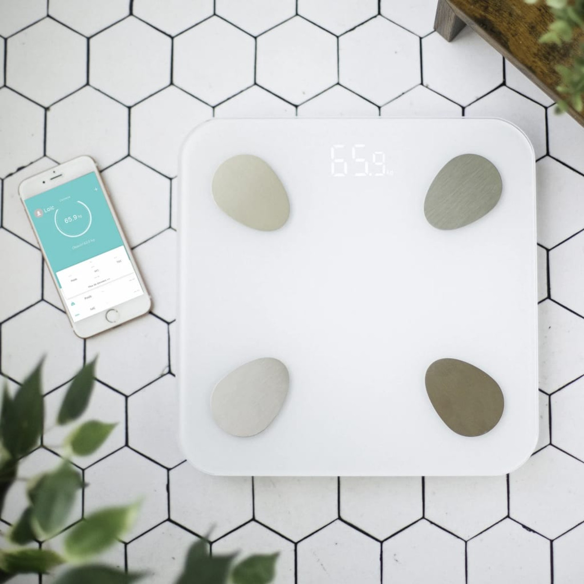 Biała waga cyfrowa z czterema szarymi owalami, telefonem wyświetlającym dane i kafelkami z roślinami.
