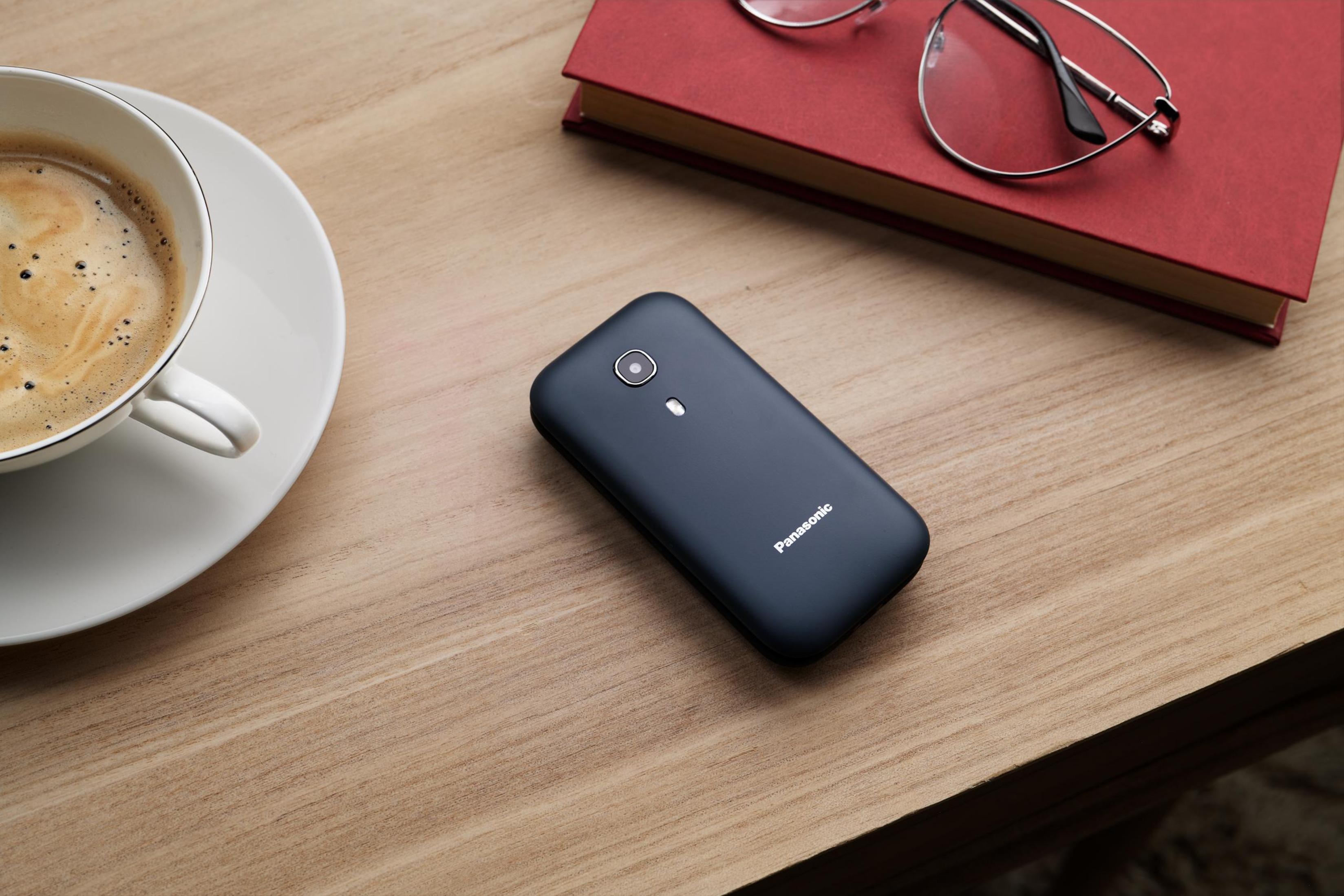 Ein schwarzes Telefon auf einem Holztisch neben einer Kaffeetasse, einem Buch und einer Brille.