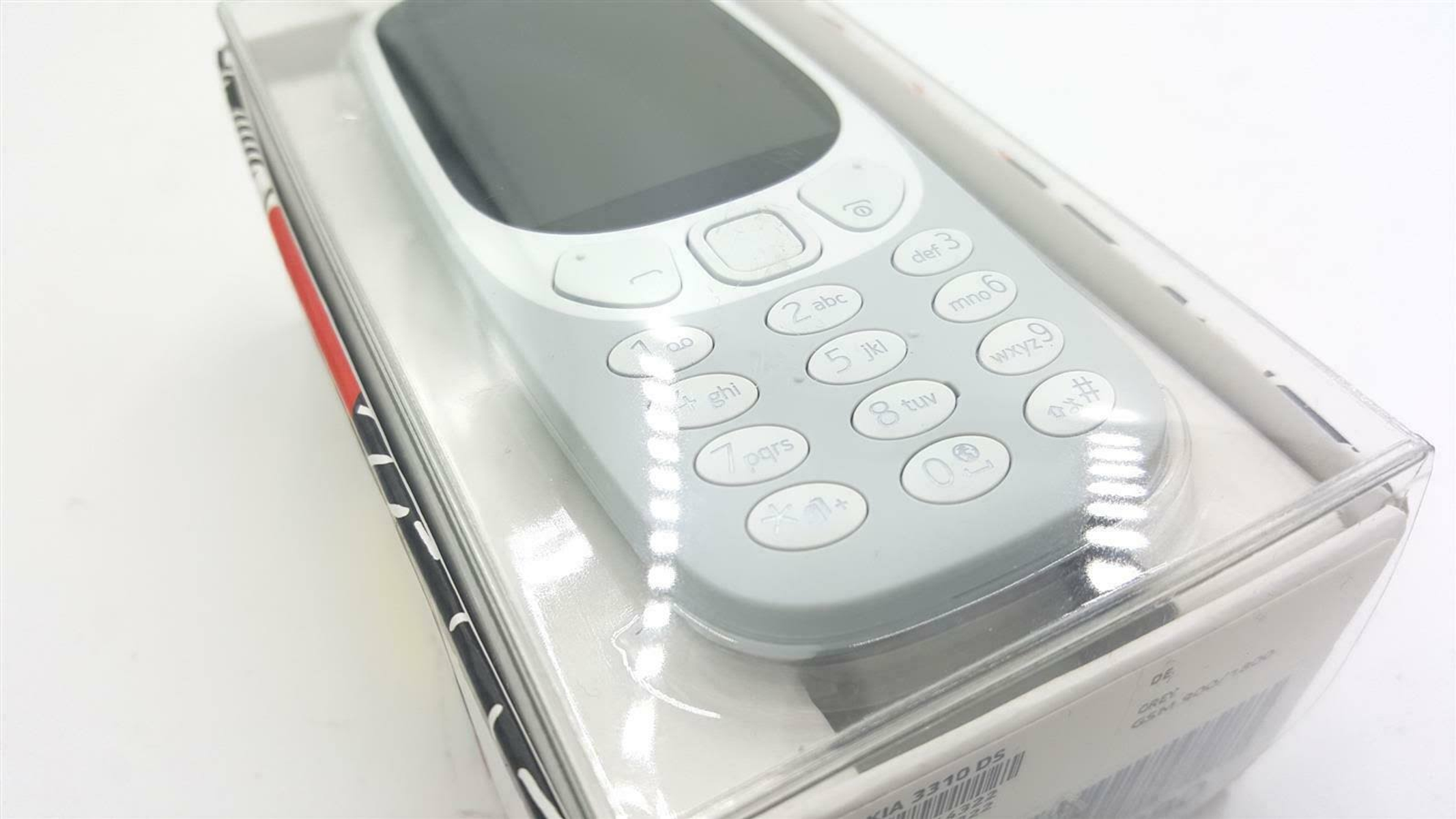 Jasnoszary telefon w przezroczystym plastikowym pudełku. Białe tło.