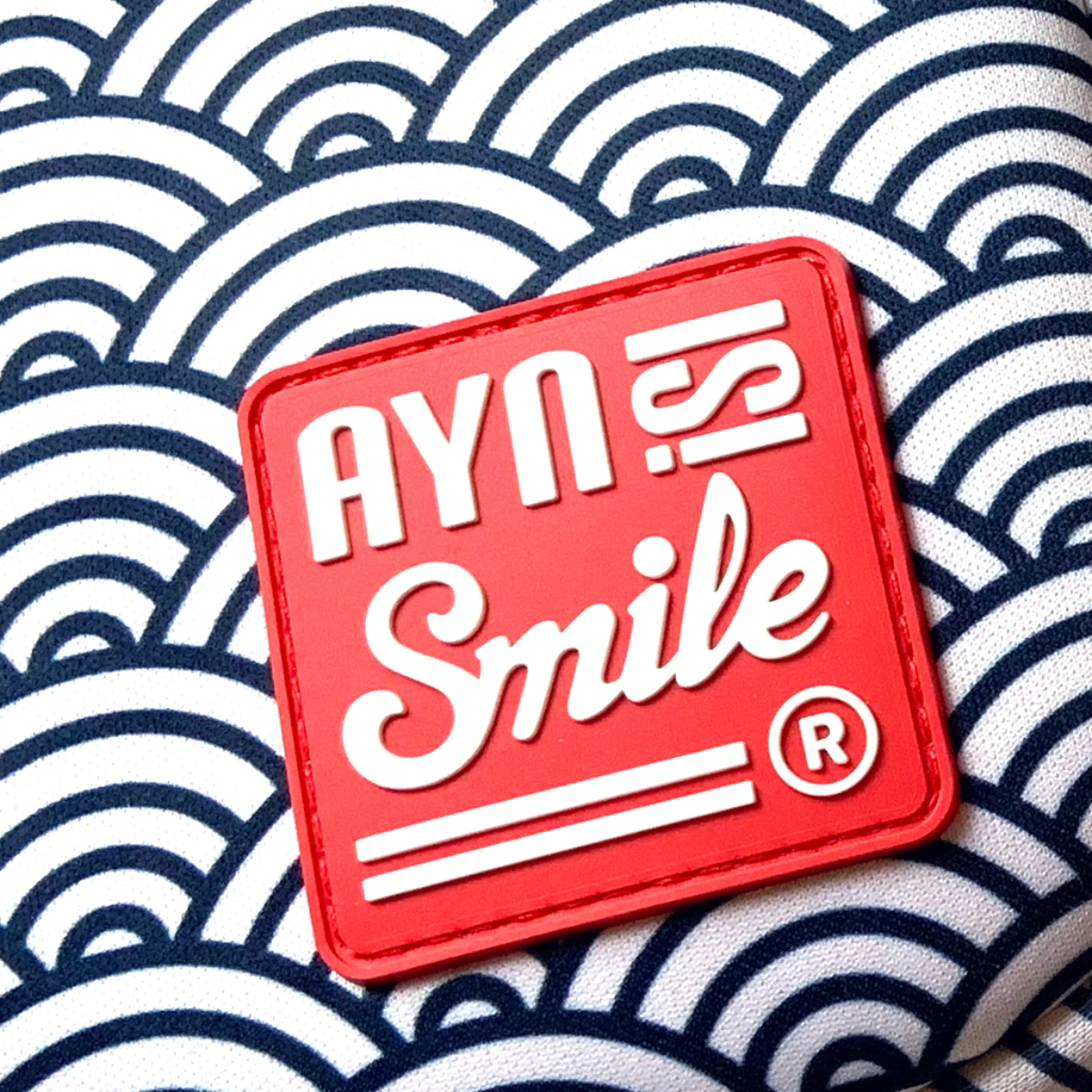 Cuadrado rojo con texto blanco: AYN IS! Smile. Fondo estampado.