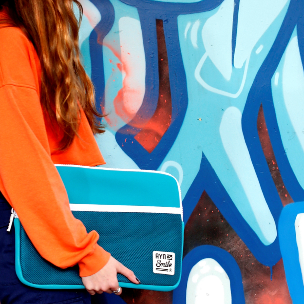 Mujer sosteniendo una funda para portátil azul y verde azulado frente a una pared de graffiti.