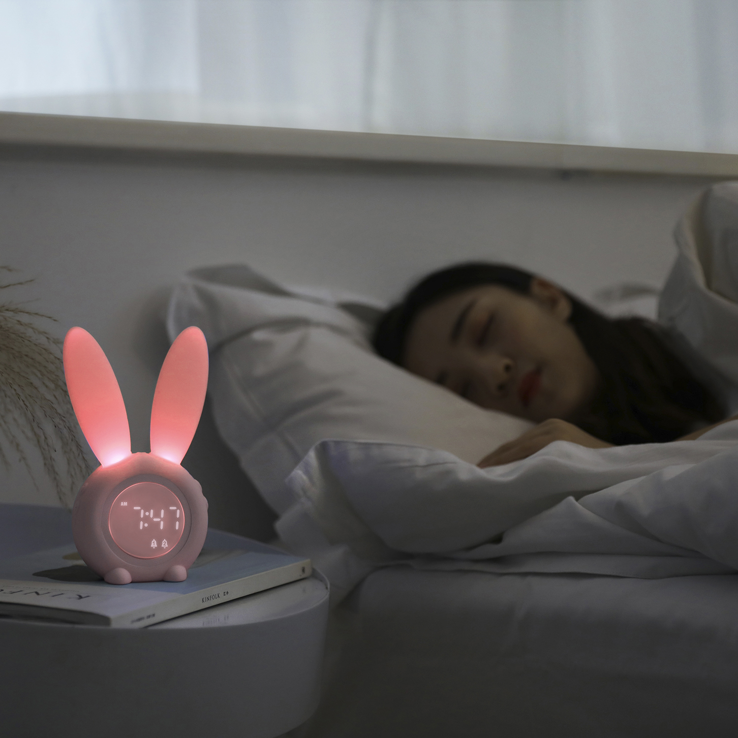 Un despertador rosa con forma de conejo en una mesita de noche. Una mujer duerme al fondo.