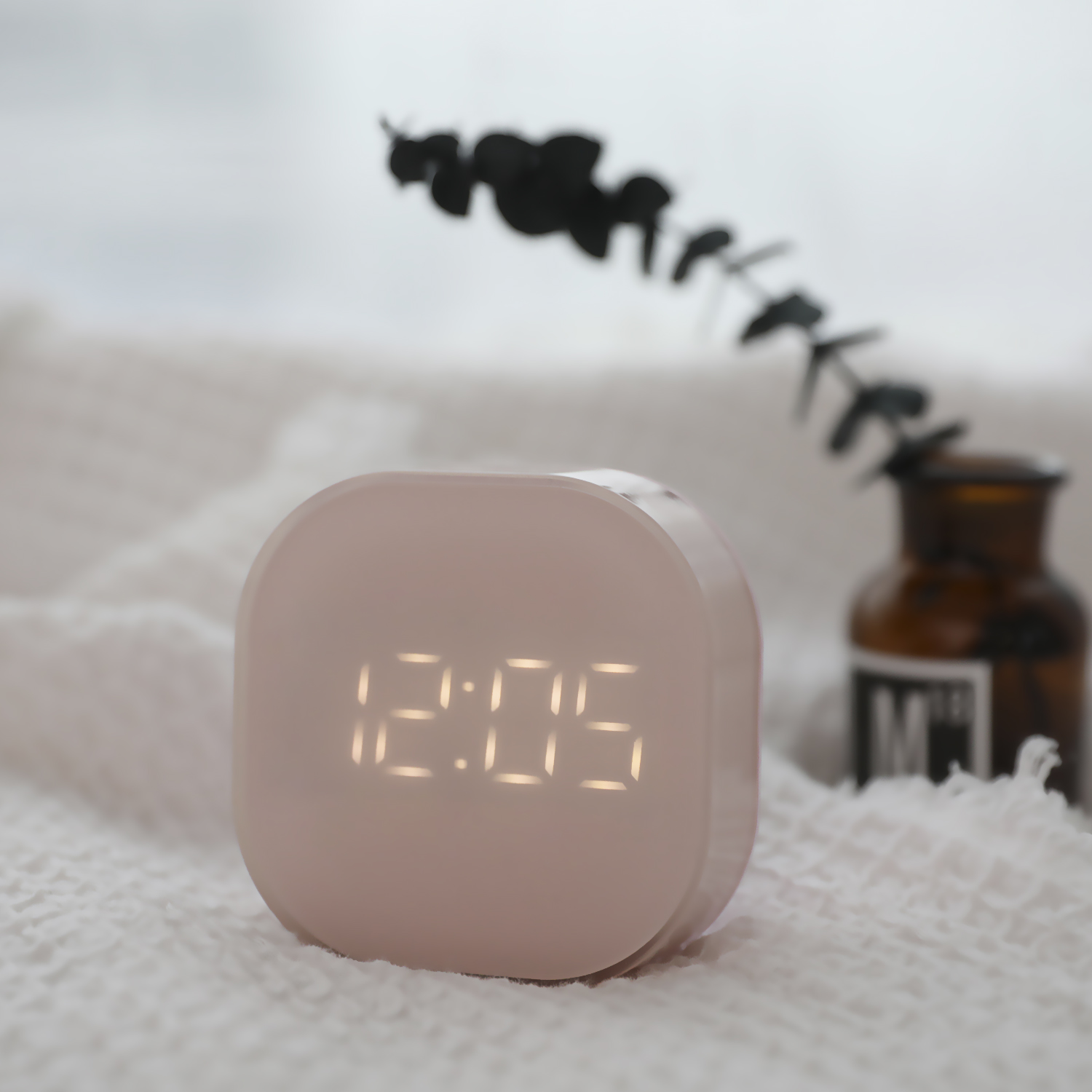 Un pequeño reloj digital rosa que muestra 12:05. Está sobre una superficie blanca con fondo borroso.