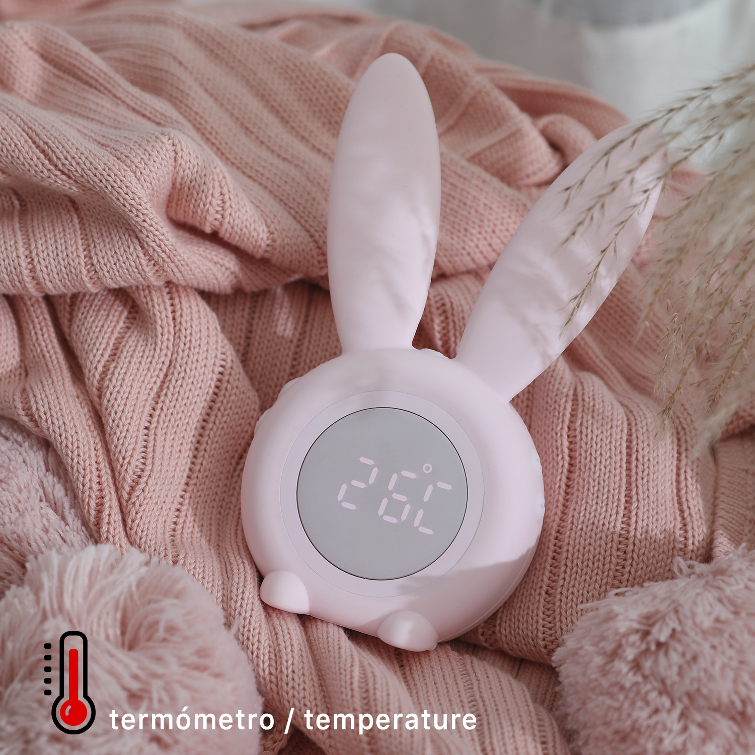 Reloj rosa con forma de conejito con pantalla digital, etiqueta de termómetro y manta rosa.