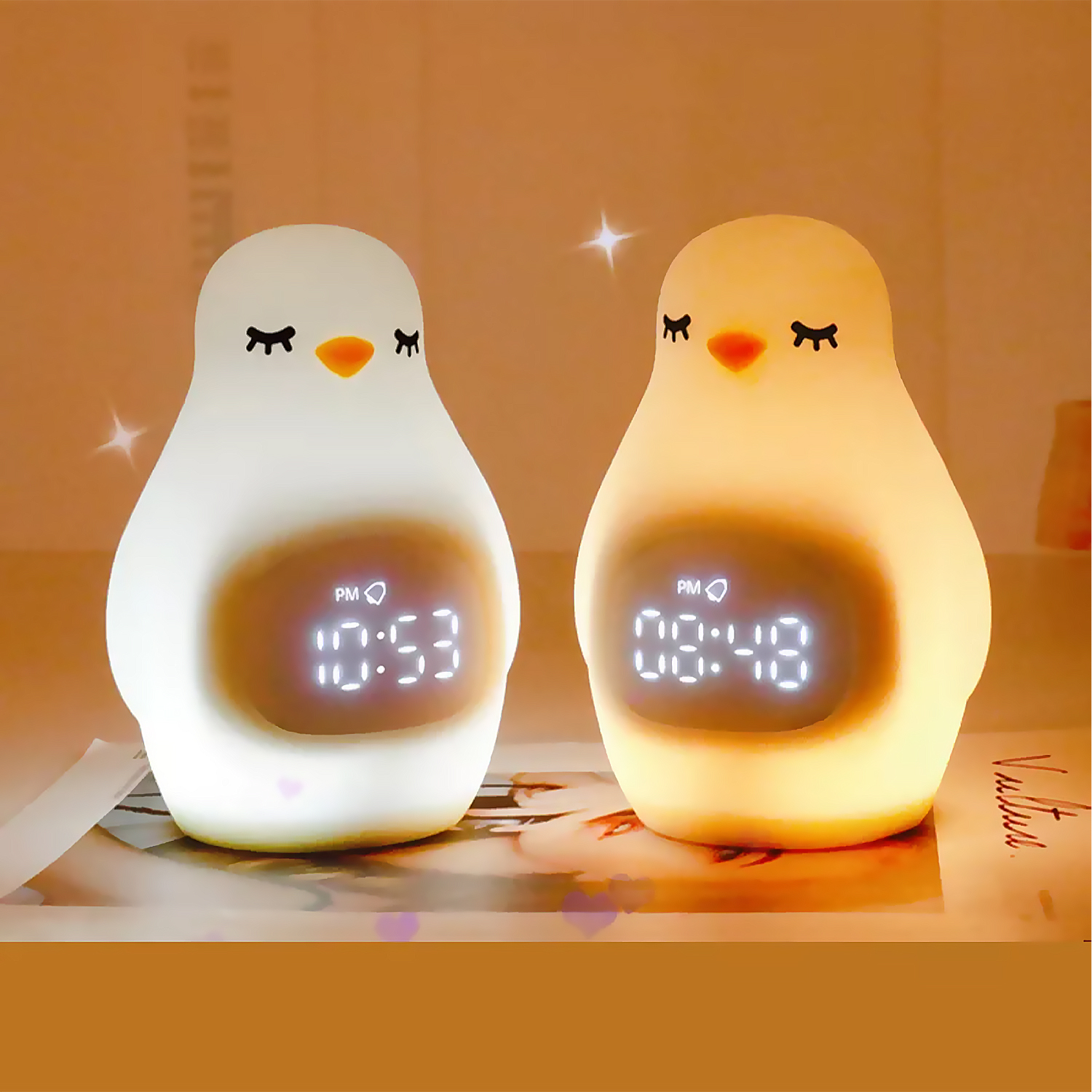 Dos luces de noche con forma de pingüino con relojes digitales que muestran diferentes horas, sobre una superficie.