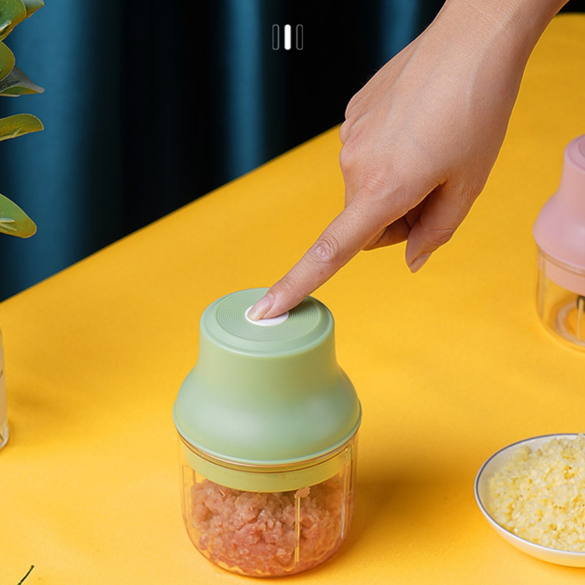 Eine Hand drückt auf einen Knopf eines grünen und durchsichtigen Foodprozessors, mit gehackten Lebensmitteln im Behälter.