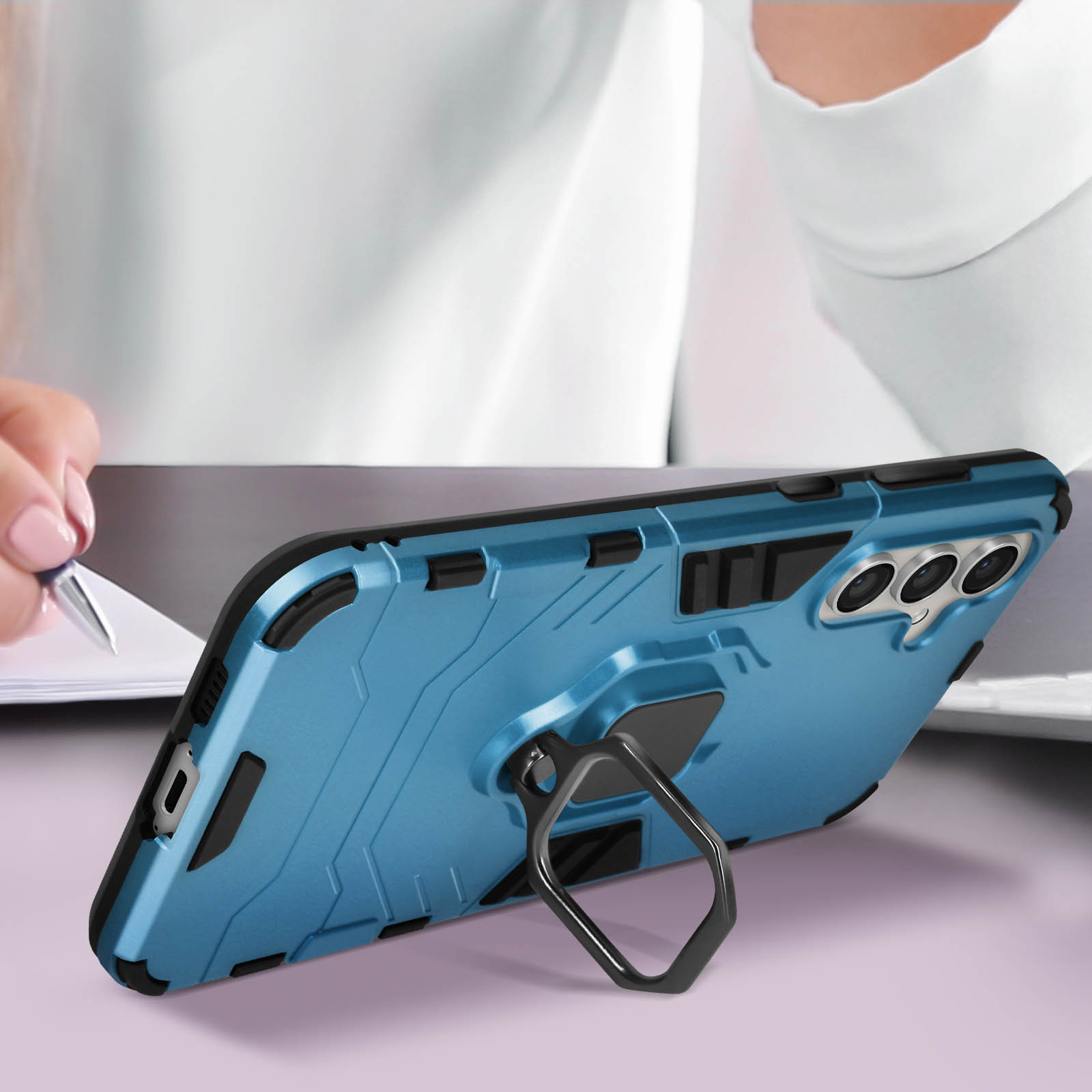 Custodia blu per telefono con supporto ad anello, su tavolo, mani di una persona che scrive su carta sullo sfondo.