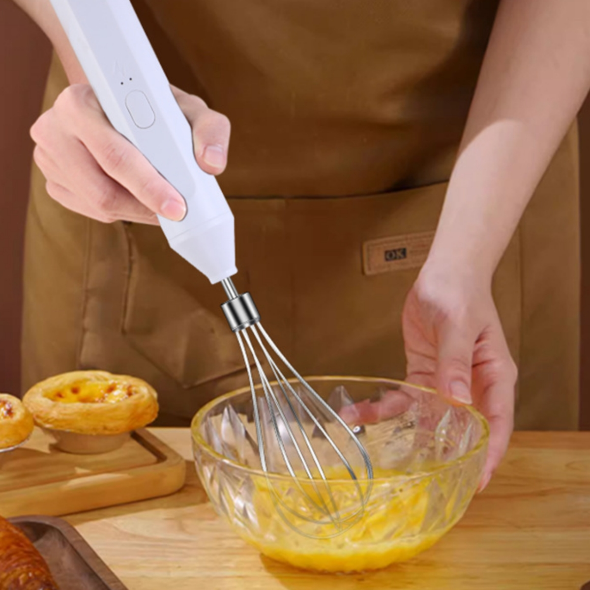 Eine Person verquirlt Eier in einer Glasschüssel mit einem weißen Handmixer, Zutaten auf einem Holztisch.