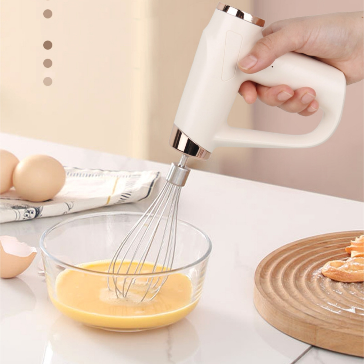 Hand hält weißen elektrischen Mixer über einer Schüssel mit geschlagenem Ei. Eier und Schneidebrett mit Essen sichtbar.