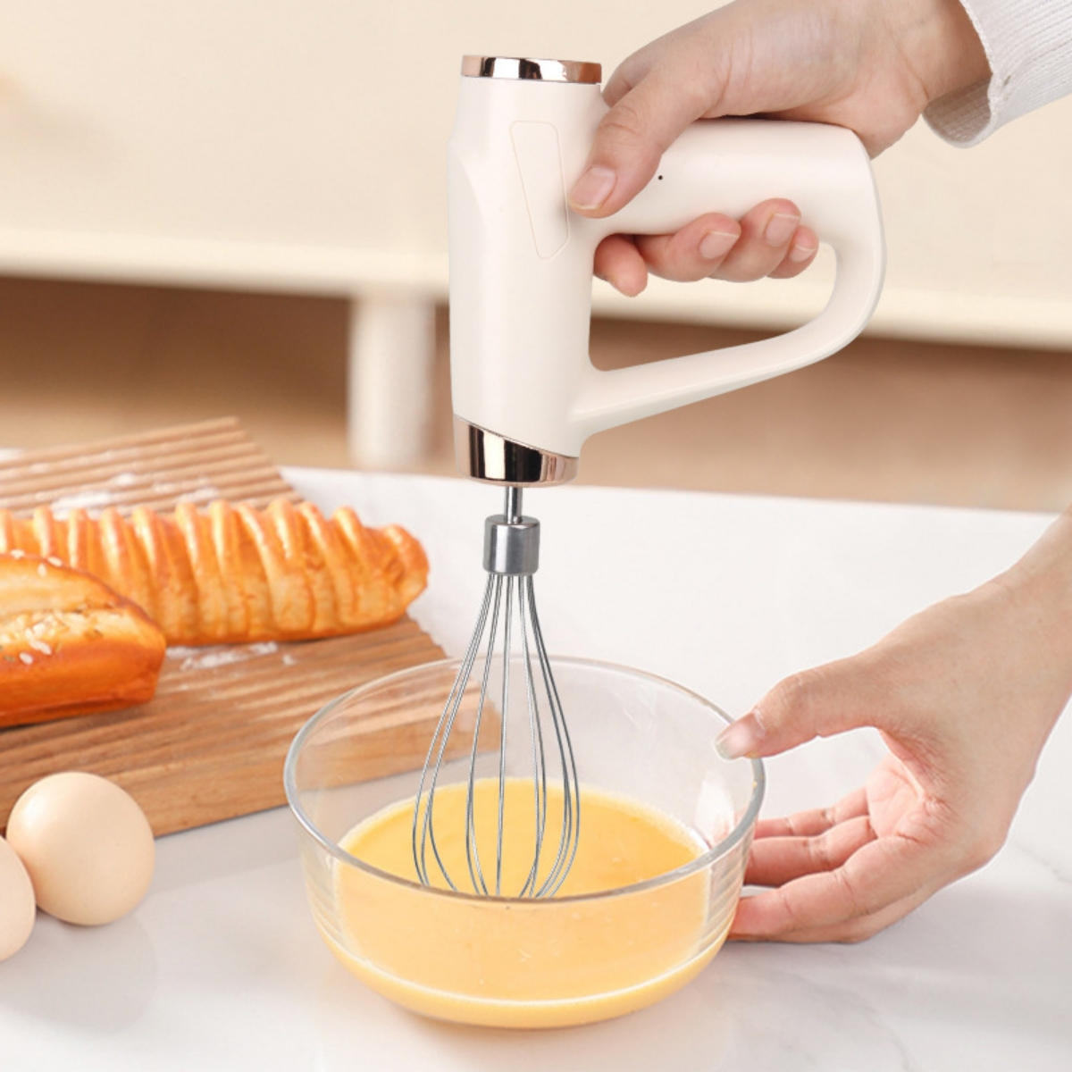 Eine Hand, die einen weißen elektrischen Mixer über einer Glasschüssel mit gelber Flüssigkeit, Eiern und Brot hält.