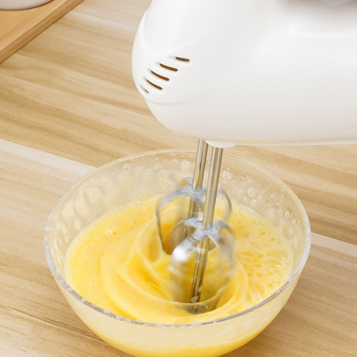 Ein weißer Handmixer verrührt Eier in einer Glasschüssel. Die Schüssel steht auf einer Holzoberfläche.