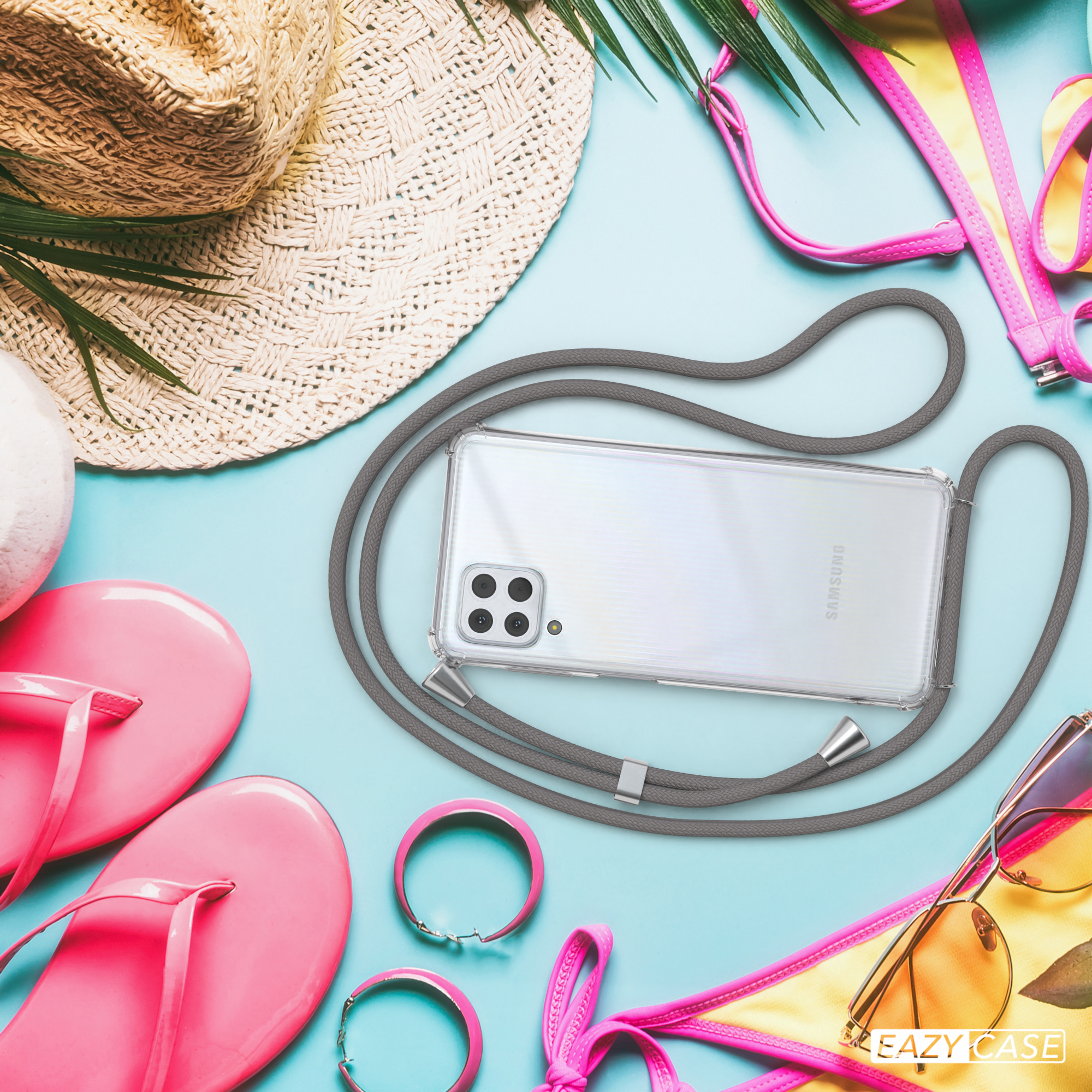 Ein Telefon mit Hülle und Riemen liegt neben Strandartikeln: Hut, Sonnenbrille und rosa Accessoires.