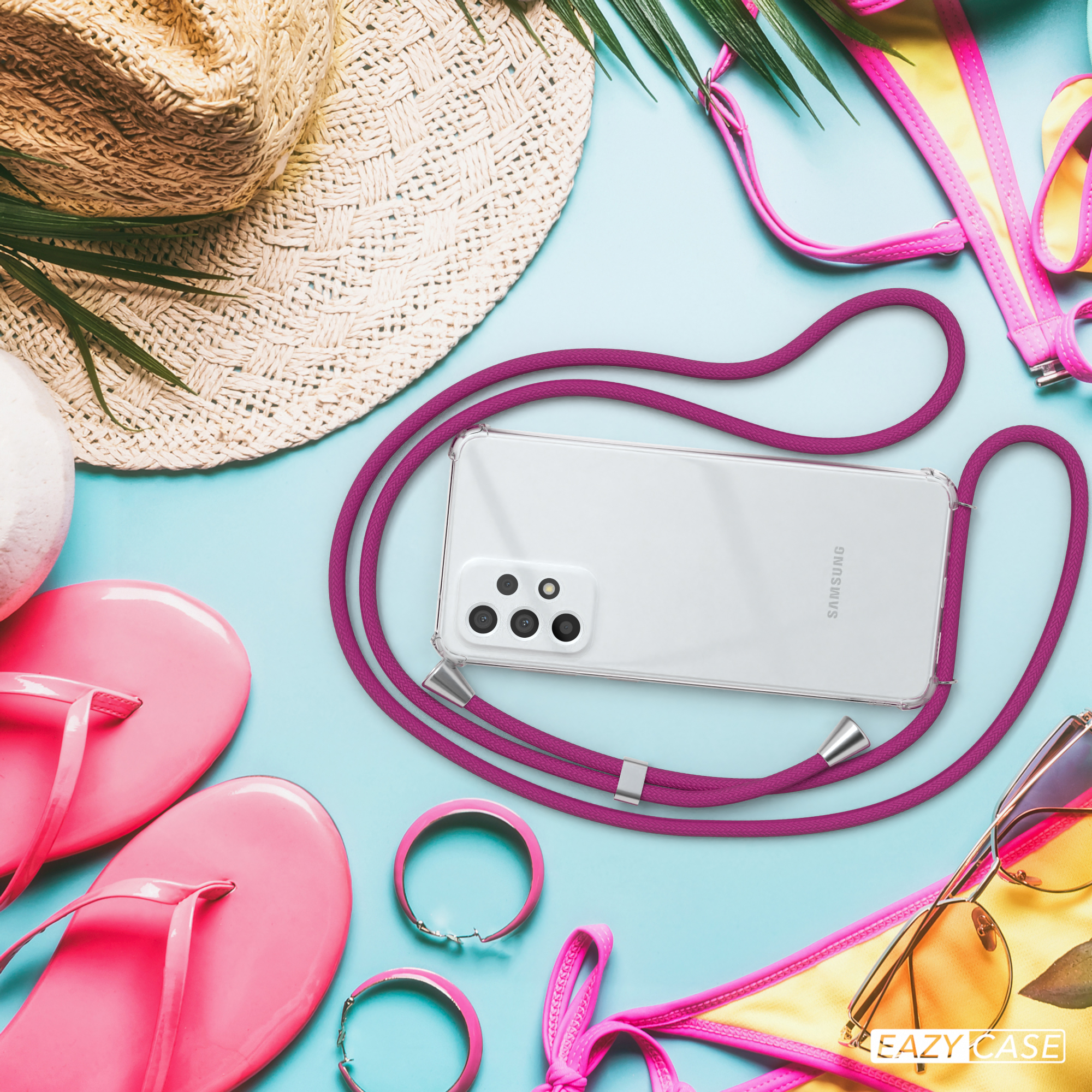 Ein Handy mit pinkfarbenem Umhängeband liegt am Strand. Sonnenbrille, Flip-Flops und Hut sind vorhanden.