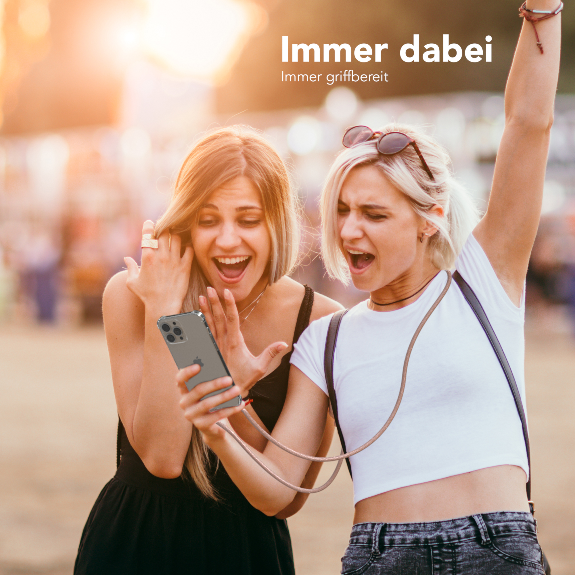 Zwei Frauen betrachten aufgeregt ein Telefon, eine mit erhobenem Arm. Sonnenuntergang im Hintergrund.