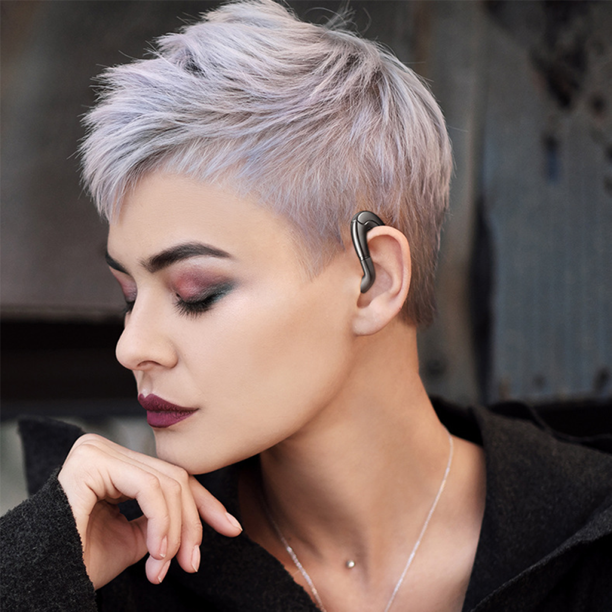 Eine Frau mit kurzen, grauen Haaren trägt einen schwarzen Ohrhörer. Sie stützt ihr Kinn auf ihre Hand und trägt eine dunkle Jacke und Halskette.