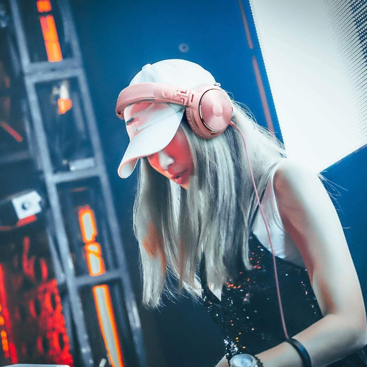 Eine Frau DJ mit Kopfhörern und Mütze, in einem Nachtclub mit blauer Beleuchtung. Sie schaut nach unten.