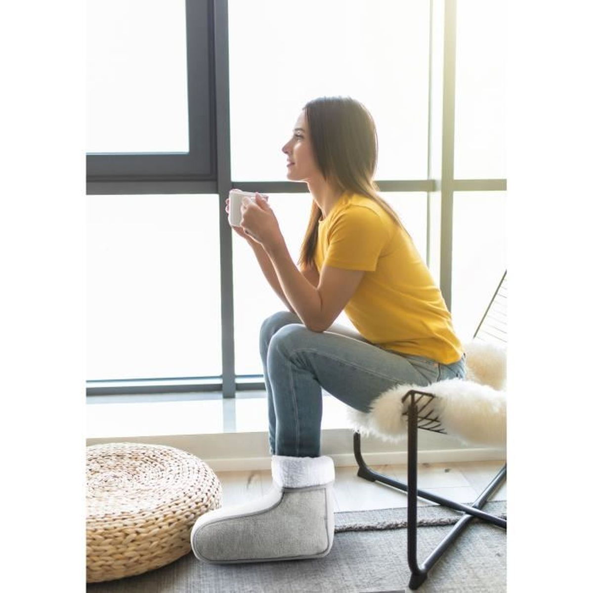 Una donna seduta su una sedia, con una tazza in mano, indossa una camicia gialla, jeans e stivali grigi.