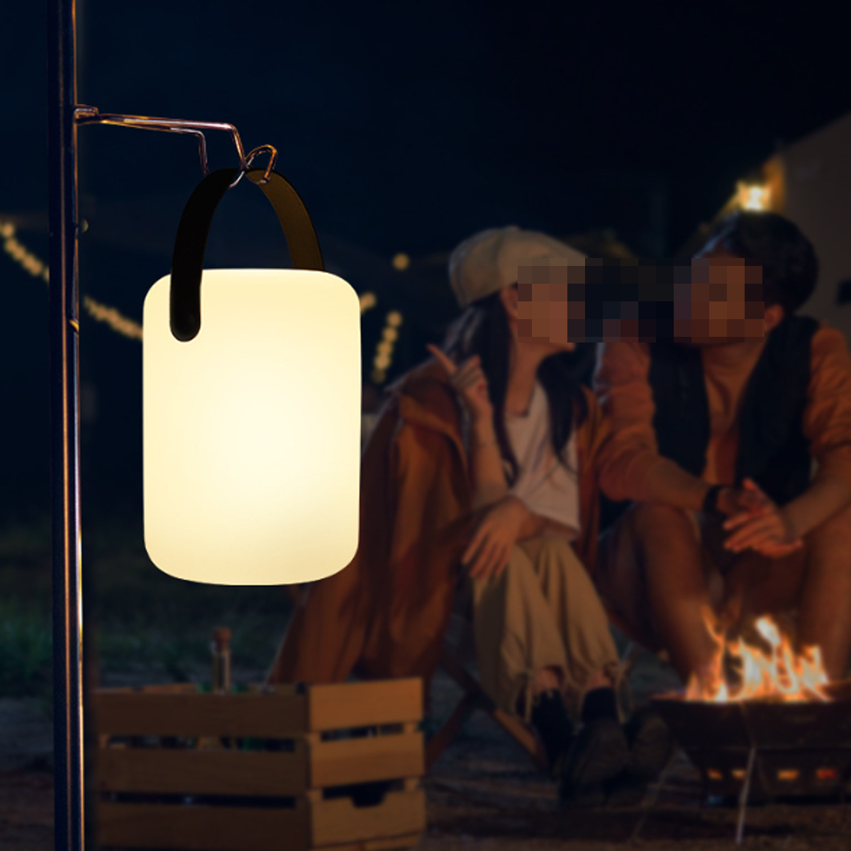 Eine leuchtende Laterne hängt in der Nähe eines Lagerfeuers, mit verschwommenen Personen im Hintergrund bei Nacht.