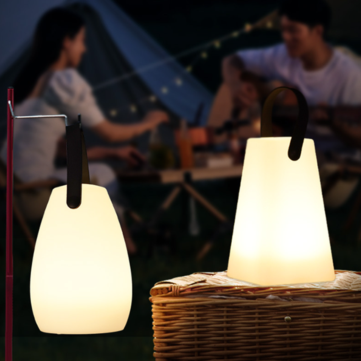 Zwei weiße, leuchtende Lampen auf einem Picknickkorb, im Hintergrund ein Paar auf einem Campingplatz.