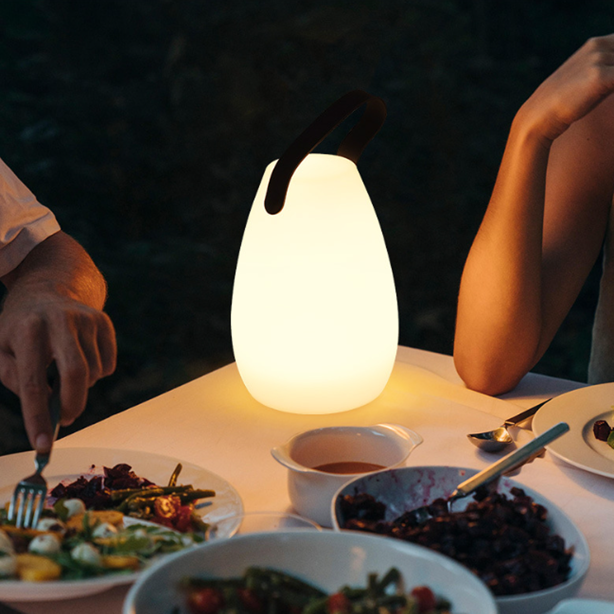 Eine leuchtende weiße Lampe auf einem Tisch mit Essen, neben Armen von Personen im Dunkeln.